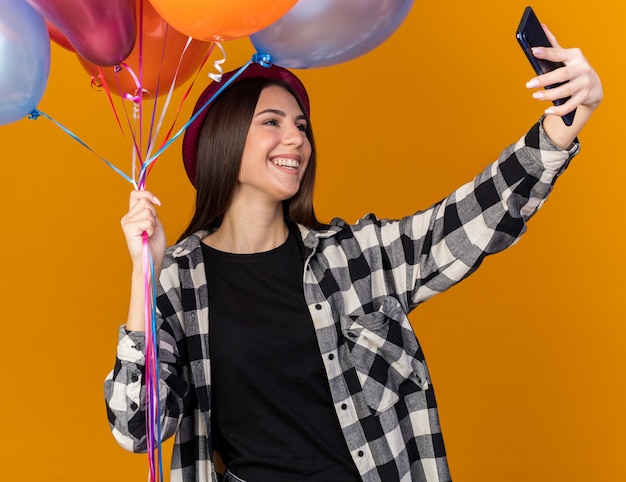 Smiling young beautiful girl wearing party hat holding balloons and take a selfie isolated on orange wall