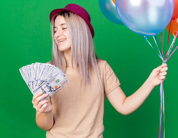 Smiling young beautiful girl wearing party hat with braces holding balloons holding out cash at camera 