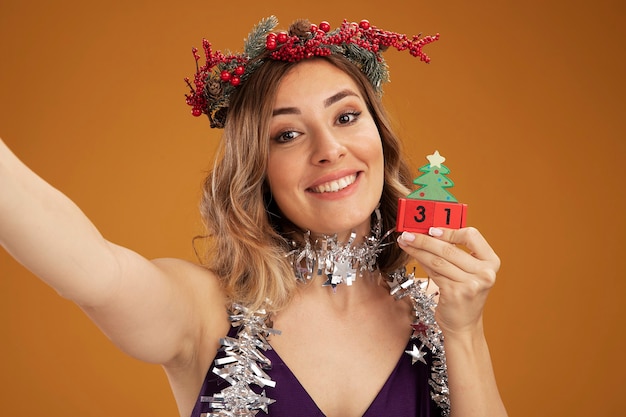 Smiling young beautiful girl wearing purple dress and wreath with garland on neck holding christmas toy and camera isolated on brown background