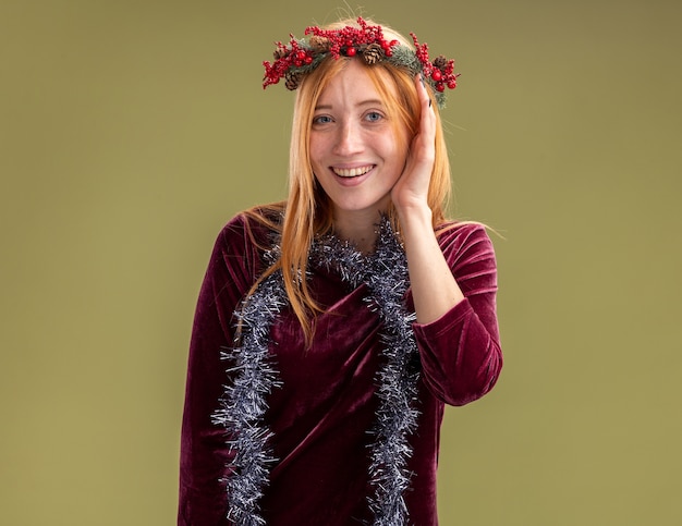 Free photo smiling young beautiful girl wearing red dress with wreath and garland on neck covered face with hand isolated on olive green background