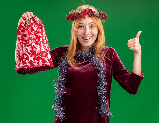 Smiling young beautiful girl wearing red dress with wreath and garland on neck holding christmas bag showing thumb up isolated on green background