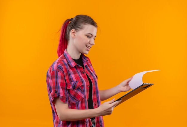 Smiling young beautiful girl wearing red shirt flipping through clipboard in her hand on isolated yellow background