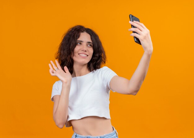 Smiling young beautiful woman holding mobile phone and waving at it on isolated orange wall with copy space