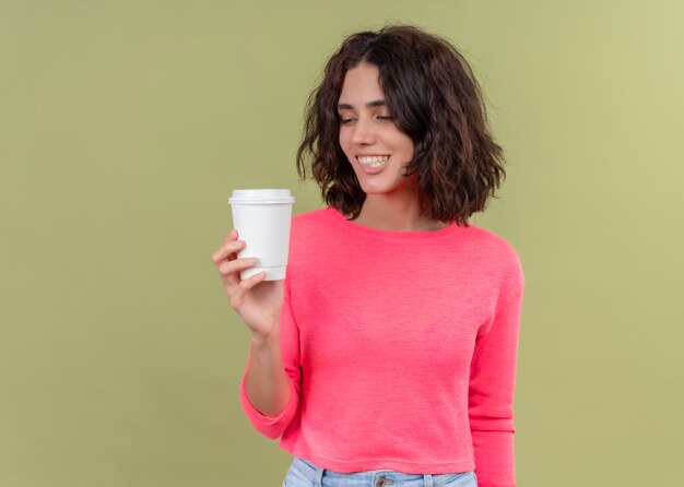 Smiling young beautiful woman holding plastic coffee cup and looking at it on isolated green wall with copy space