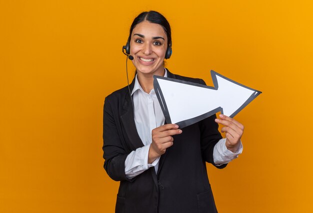 Smiling young beautiful woman wearing black blazer with headset holding direction mark 