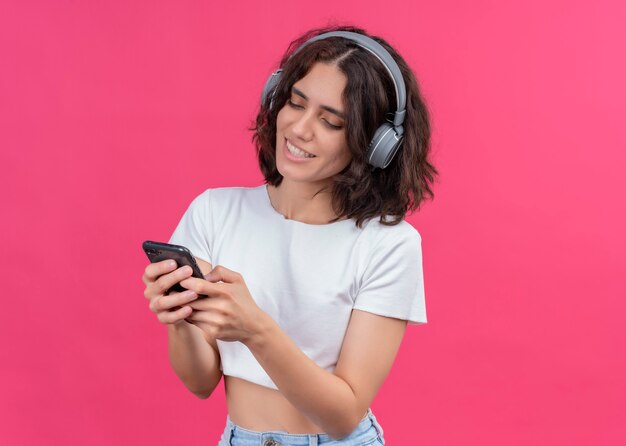 Smiling young beautiful woman wearing headphones and holding mobile phone on pink wall with copy space