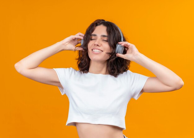 Smiling young beautiful woman wearing headphones and putting hands on them with closed eyes on isolated orange wall