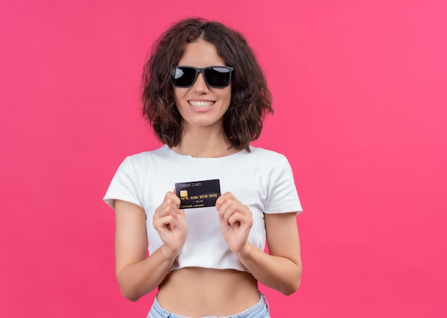 Smiling young beautiful woman wearing sunglasses and holding card on isolated pink wall with copy space