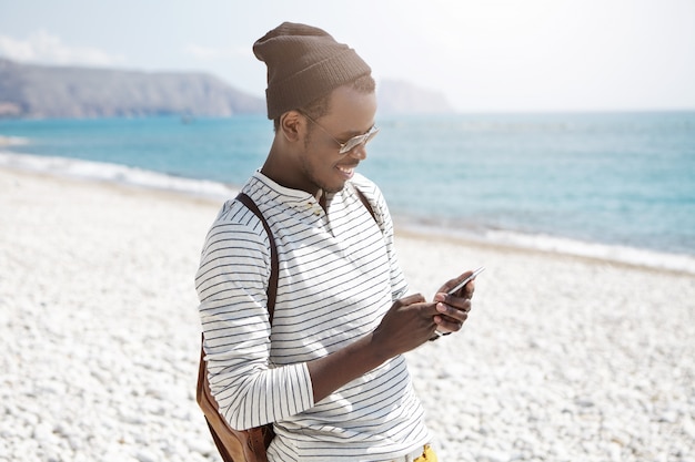 Smiling young black European male tourist in hat and shades using 3g internet on mobile phone on beach, sharing pictures via social media, enjoying happy days during his summer vacations at seaside