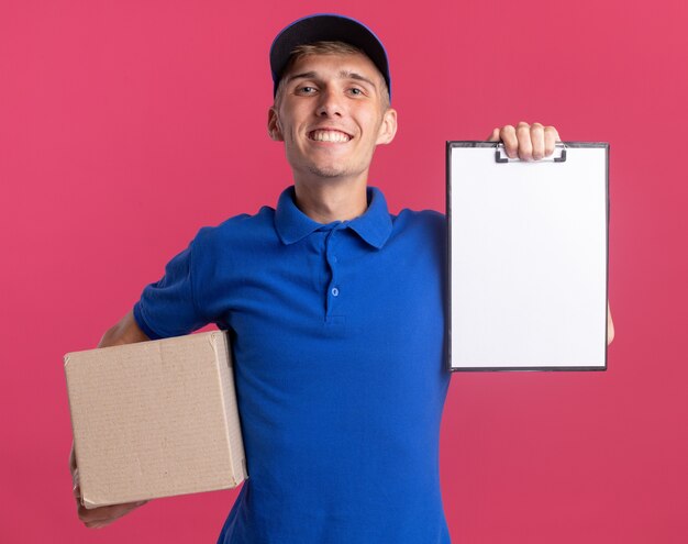 Smiling young blonde delivery boy holding clipboard and cardbox