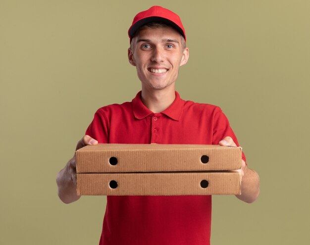 Smiling young blonde delivery boy holding pizza boxes on olive green 