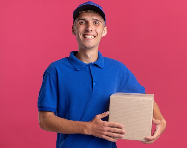 Smiling young blonde delivery boy holds cardbox isolated on pink wall with copy space