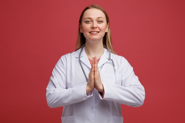 Smiling young blonde female doctor wearing medical robe and stethoscope around neck doing namaste gesture 