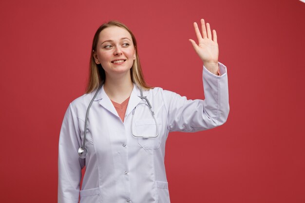 Smiling young blonde female doctor wearing medical robe and stethoscope around neck looking at side waving 