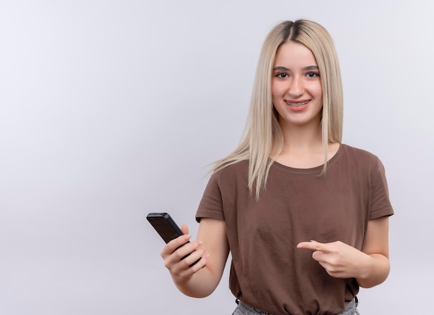 Free photo smiling young blonde girl in dental braces holding mobile phone and pointing at it on isolated white space with copy space