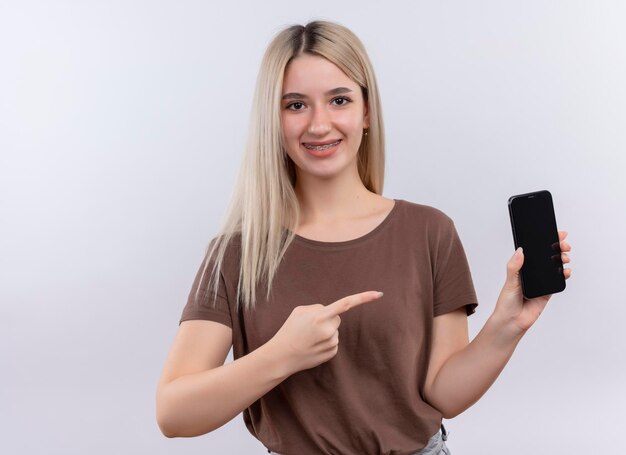 Smiling young blonde girl in dental braces holding mobile phone and pointing at it on isolated white space with copy space