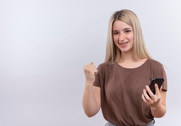 Smiling young blonde girl in dental braces holding mobile phone with raised fist on isolated white space with copy space