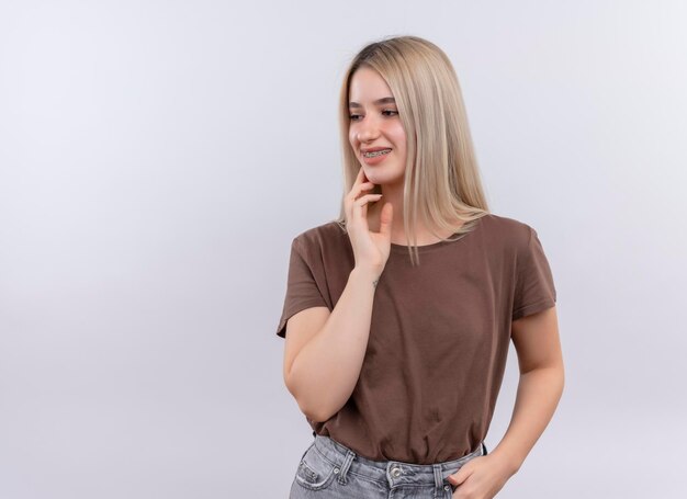 Smiling young blonde girl in dental braces with hand on chin on isolated white space with copy space