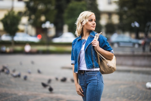 Smiling Young blonde girl woman on streetwalk square fontain dressed up in blue jeans suite with bag on her shoulder in sunny day