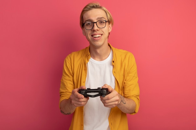 Free photo smiling young blonde guy wearing yellow t-shirt and glasses game controller joystick isolated on pink