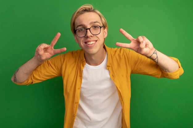 Free Photo smiling young blonde guy wearing yellow t-shirt and glasses showing peace gesture isolated on green