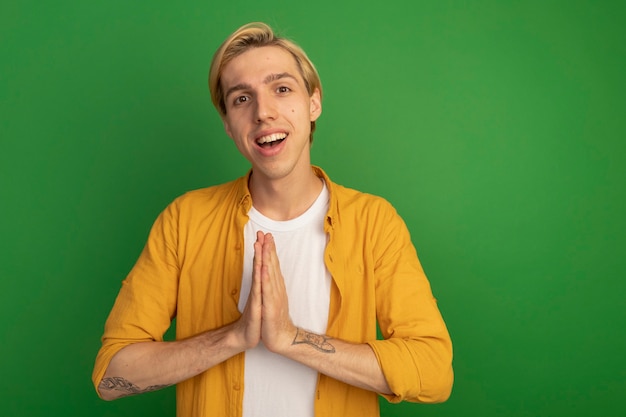 Free photo smiling young blonde guy wearing yellow t-shirt showing pray gesture isolated on green with copy space