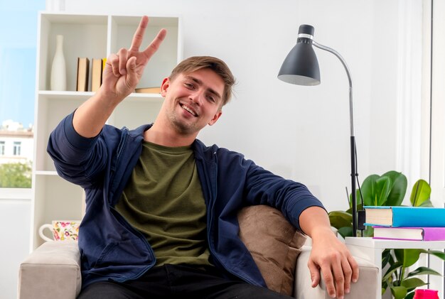 Smiling young blonde handsome man sits on armchair inside living room