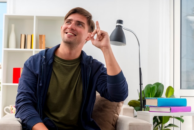 Smiling young blonde handsome man sits on armchair pointing up looking at side inside living room