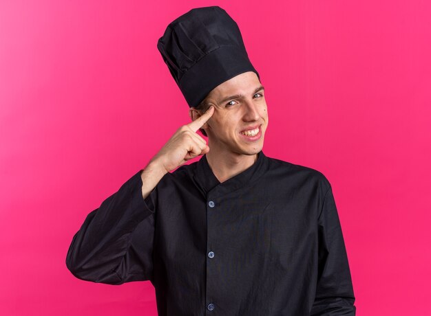 Smiling young blonde male cook in chef uniform and cap doing think gesture 