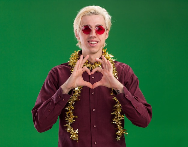 Free photo smiling young blonde man wearing glasses with tinsel garland around neck looking at camera doing heart sign isolated on green background