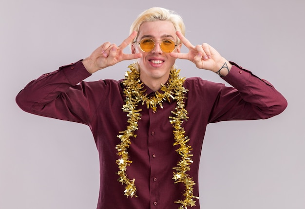 Free photo smiling young blonde man wearing glasses with tinsel garland around neck  showing v-sign symbols near eyes isolated on white wall