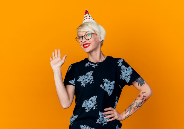 Free photo smiling young blonde party girl wearing glasses and birthday cap looking at camera keeping hand on waist doing hi gesture camera isolated on orange background with copy space