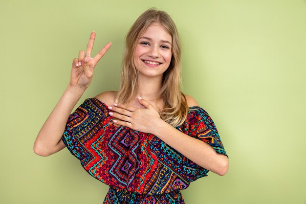 Smiling young blonde slavic girl putting hand on her chest and gesturing victory sign