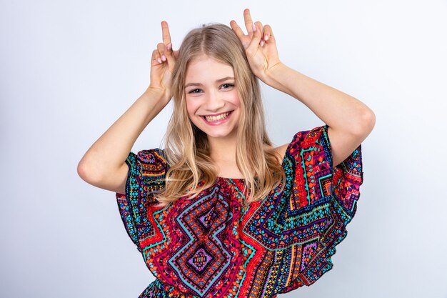 Smiling young blonde slavic girl putting hands on her head and gesturing horns with fingers