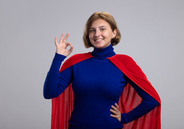 Smiling young blonde superhero girl in red cape keeping hand on waist looking at camera doing ok sign isolated on white background