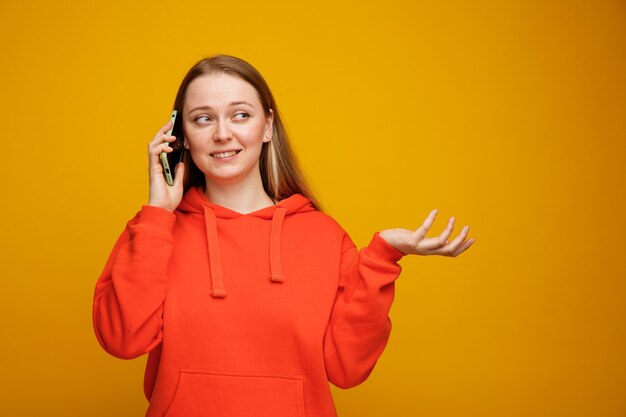 Smiling young blonde woman talking on phone looking at side showing empty hand 