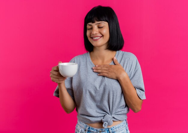 Smiling young brunette caucasian girl holds cup and puts hand on chest 