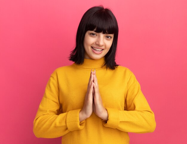 Smiling young brunette caucasian girl holds hands together on pink