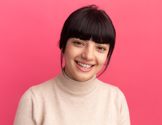 Smiling young brunette caucasian girl looks at camera isolated on pink wall with copy space