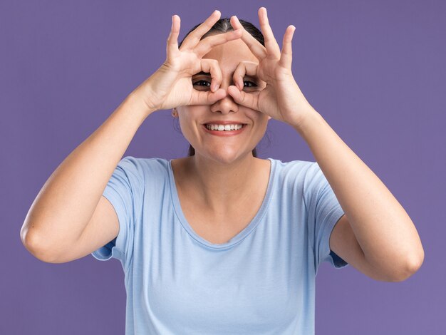 Smiling young brunette caucasian girl  through fingers isolated on purple wall with copy space