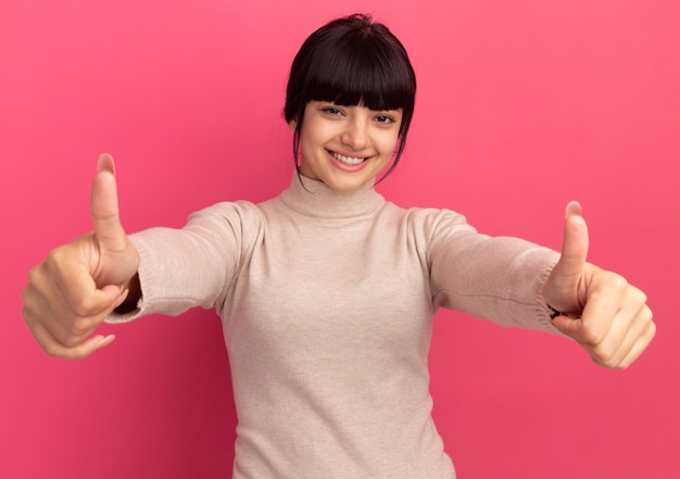 Smiling young brunette caucasian girl thumbs up with two hands on pink