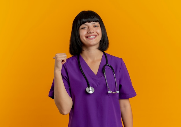 Smiling young brunette female doctor in uniform with stethoscope points behind isolated on orange background with copy space