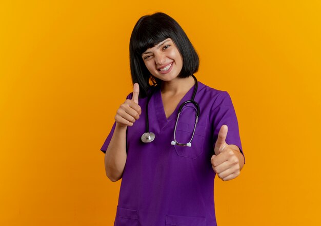 Smiling young brunette female doctor in uniform with stethoscope thumbs up with two hands isolated on orange background with copy space