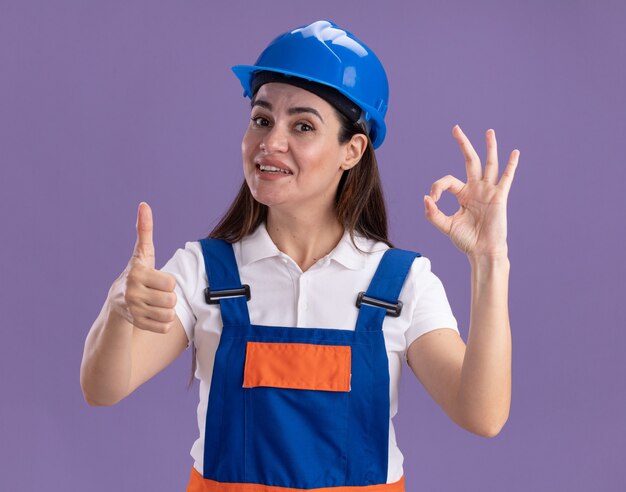 Smiling young builder woman in uniform showing okay gesture showing thumb up isolated on purple wall