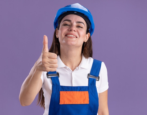 Smiling young builder woman in uniform showing thumb up isolated on purple wall