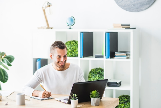 Smiling young businessman using laptop while writing on notepad