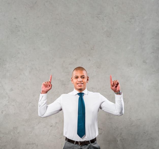 Smiling young businessman with his arms raised pointing his finger upward against grey wall