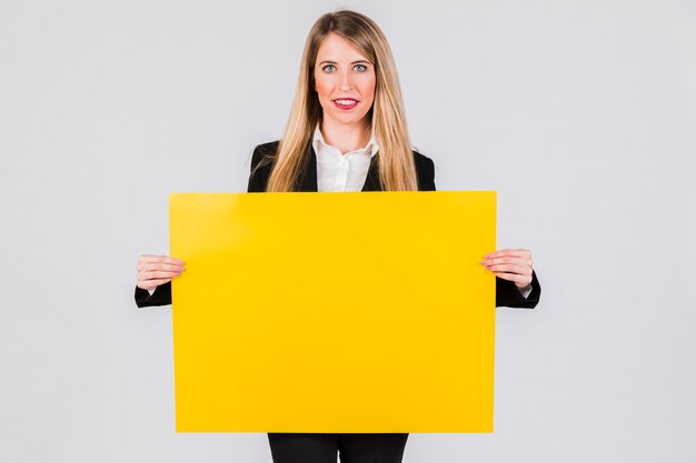 Smiling young businesswoman holding yellow blank placard against grey background