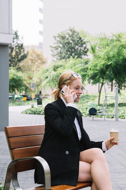 Free photo smiling young businesswoman sitting on bench holding takeaway coffee cup talking on cell phone