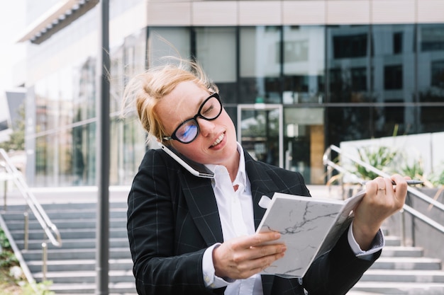 Smiling young businesswoman talking on cell phone holding diary in hand